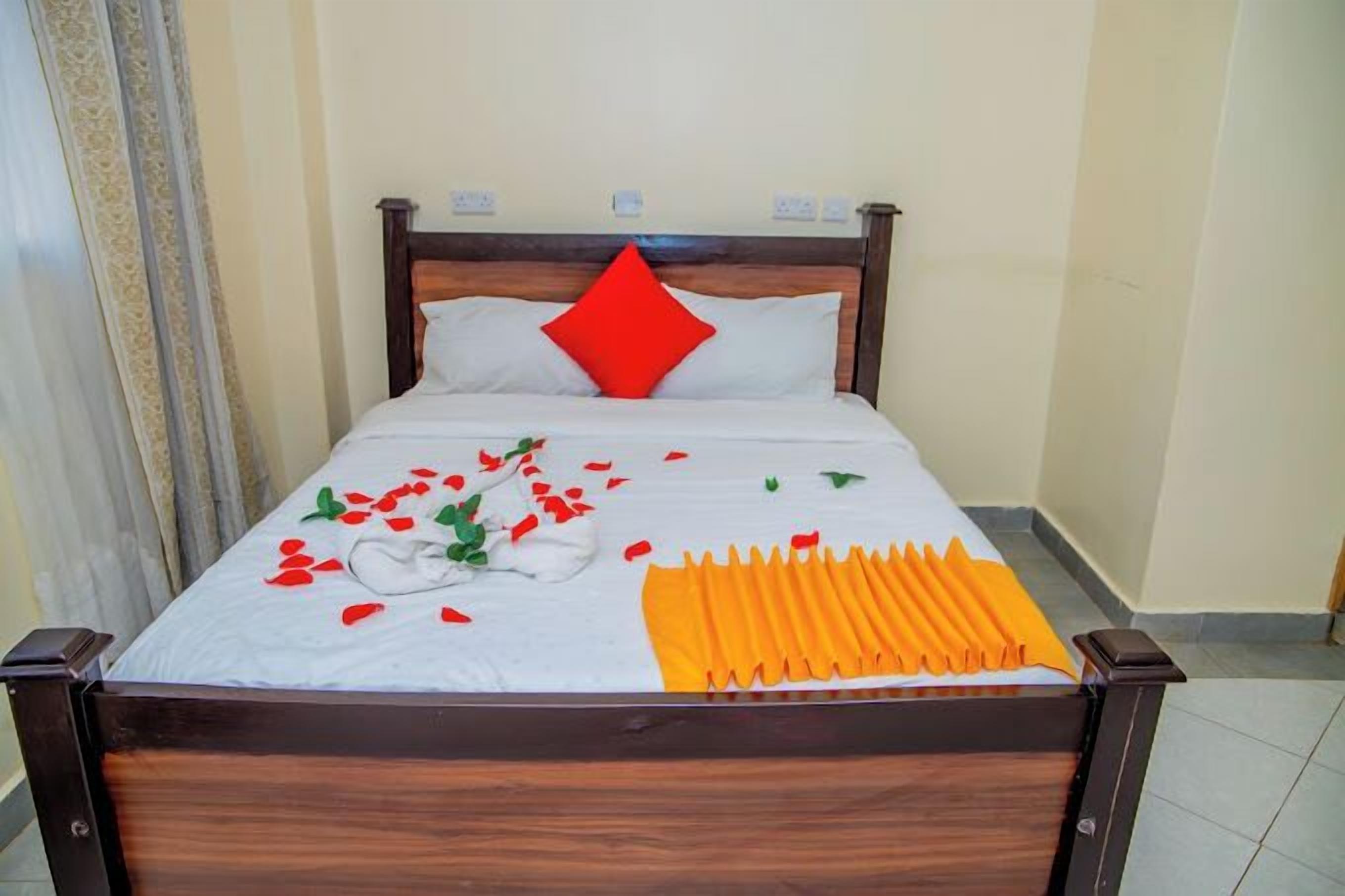 Una cama bien hecha con una colcha blanca y naranja, un cojín rojo y pétalos de rosa esparcidos.