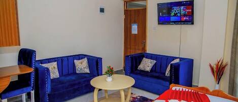Una habitación con dos sofás azules, una mesita redonda pequeña, una cama con colcha roja y blanca, y un televisor montado en la pared.