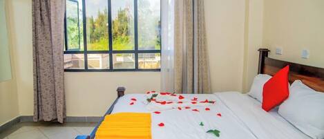 A hotel room with a large window, a bed with white bedding and red pillows, and a yellow throw blanket.