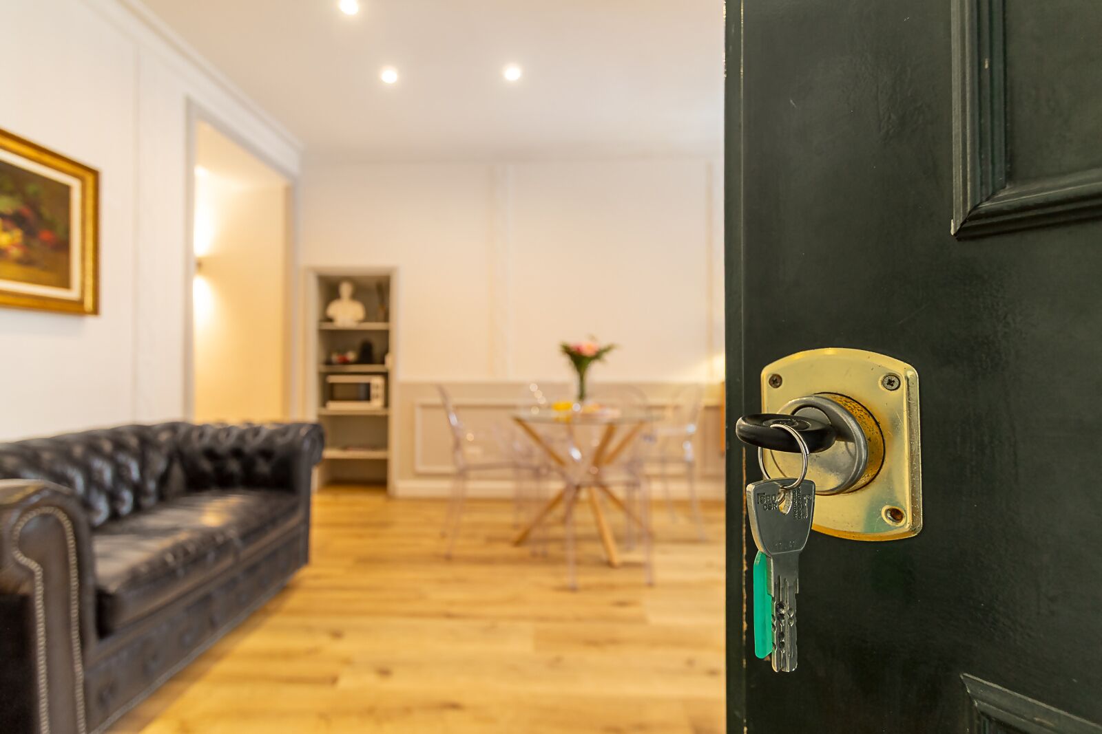 A modern living room with a black leather sofa, a dining area with a glass table, and a black door with a lock.
