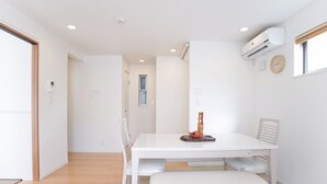 A modern, well-lit room with a white table, chairs, and a small decorative item. There are ceiling lights, an air conditioning unit, and a window with a view.