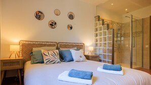 A bedroom with a large bed, two bedside tables with lamps, a glass shower enclosure, and wall art.