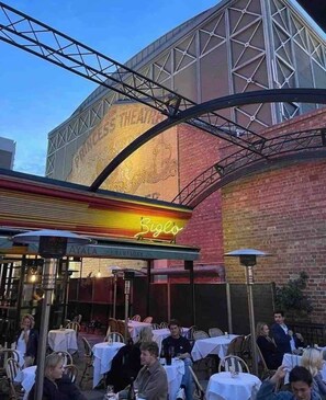 Ein Außenbereich zum Essen mit Tischen und Stühlen, einem Neonlichtschild und einem Gebäude mit einem großen Schild, auf dem 'Princess Theatre' steht.