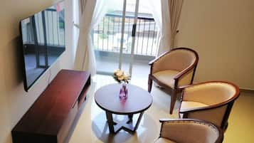 A hotel room with a TV, a wooden bench, two armchairs, a small table with a vase, and a balcony with curtains.