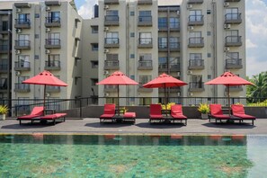 A poolside area with red umbrellas and lounge chairs, surrounded by a high-rise building with balconies. - Family-friendly 3BR Apartment in Serene Malabe (Kolonnawa)