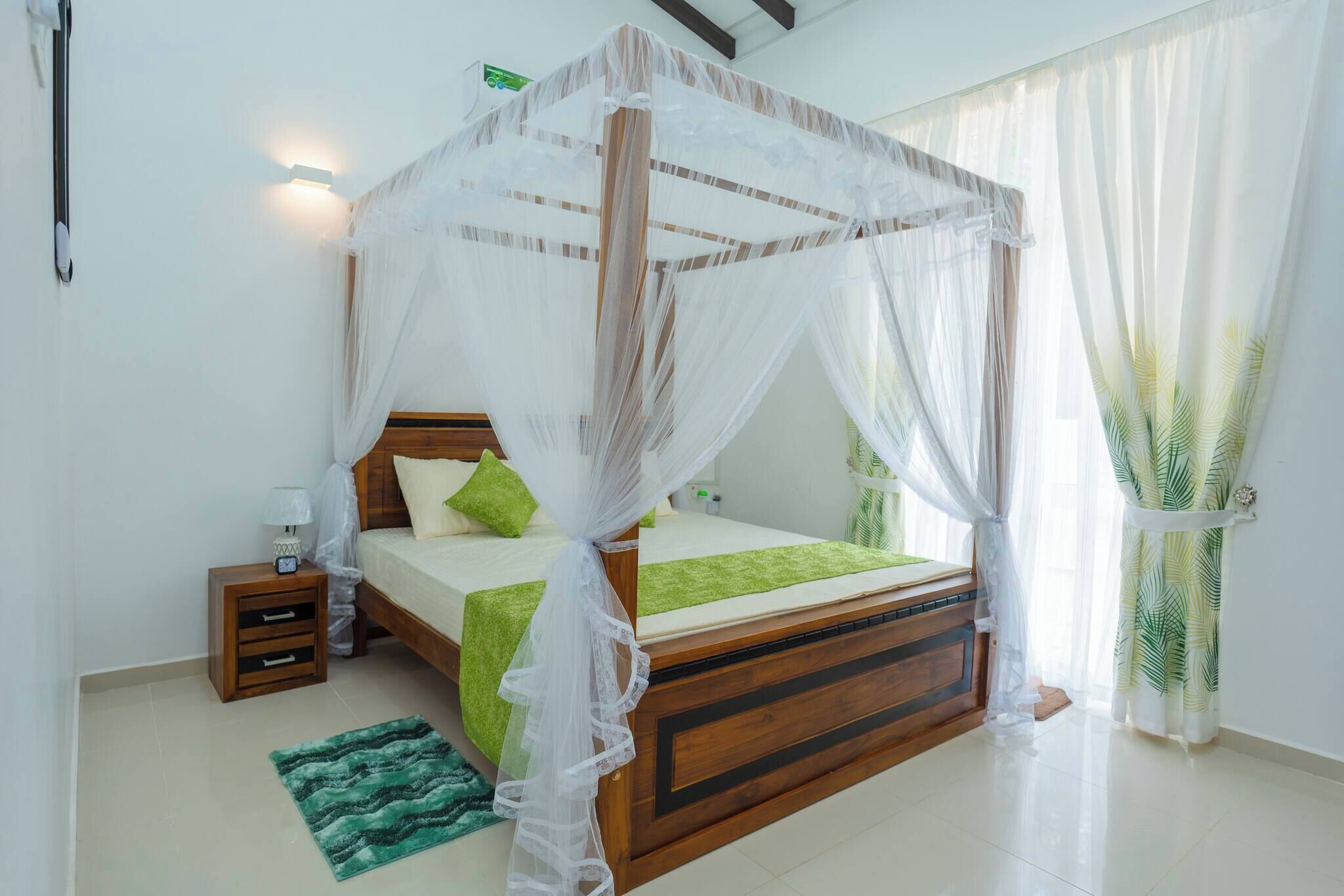 A bedroom with a wooden bed, green bedding, a nightstand, a green rug, and sheer curtains.