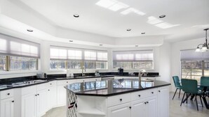 A modern kitchen with white cabinetry, a black countertop, and a central island. There are large windows providing natural light and a view of the outdoors.