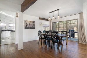 A spacious dining area with a wooden table and chairs, a large window with a view, and a modern kitchen in the background.