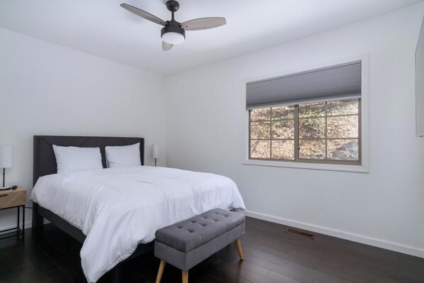 A bedroom with a bed, a nightstand, a bench, a ceiling fan, and a window with blinds. - Gallery Greens at Rumbling Bald (Lake Lure)