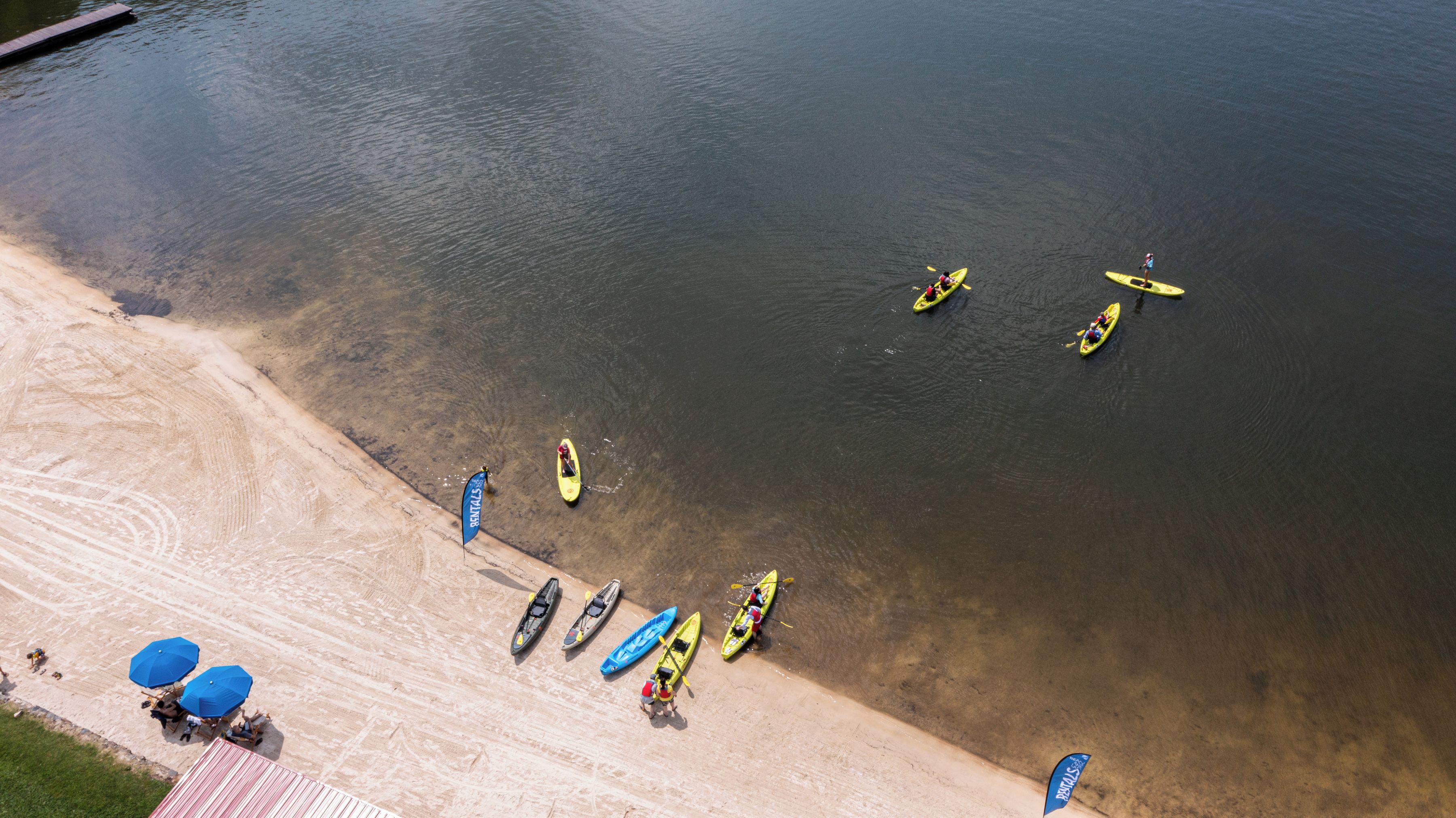 Ett flygbildsbild av en strand med kajaker uppradade nära stranden, en flagga och parasoller.