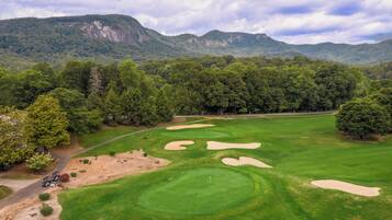 A golf course with sand bunkers and a flagstick, surrounded by dense forest and mountains.