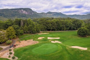 A golf course with sand bunkers and a flagstick, surrounded by dense forest and mountains.