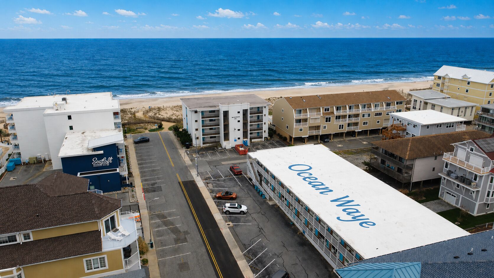 A coastal hotel with a prominent 'Ocean Way' sign, a parking lot, and a view of the beach and ocean.