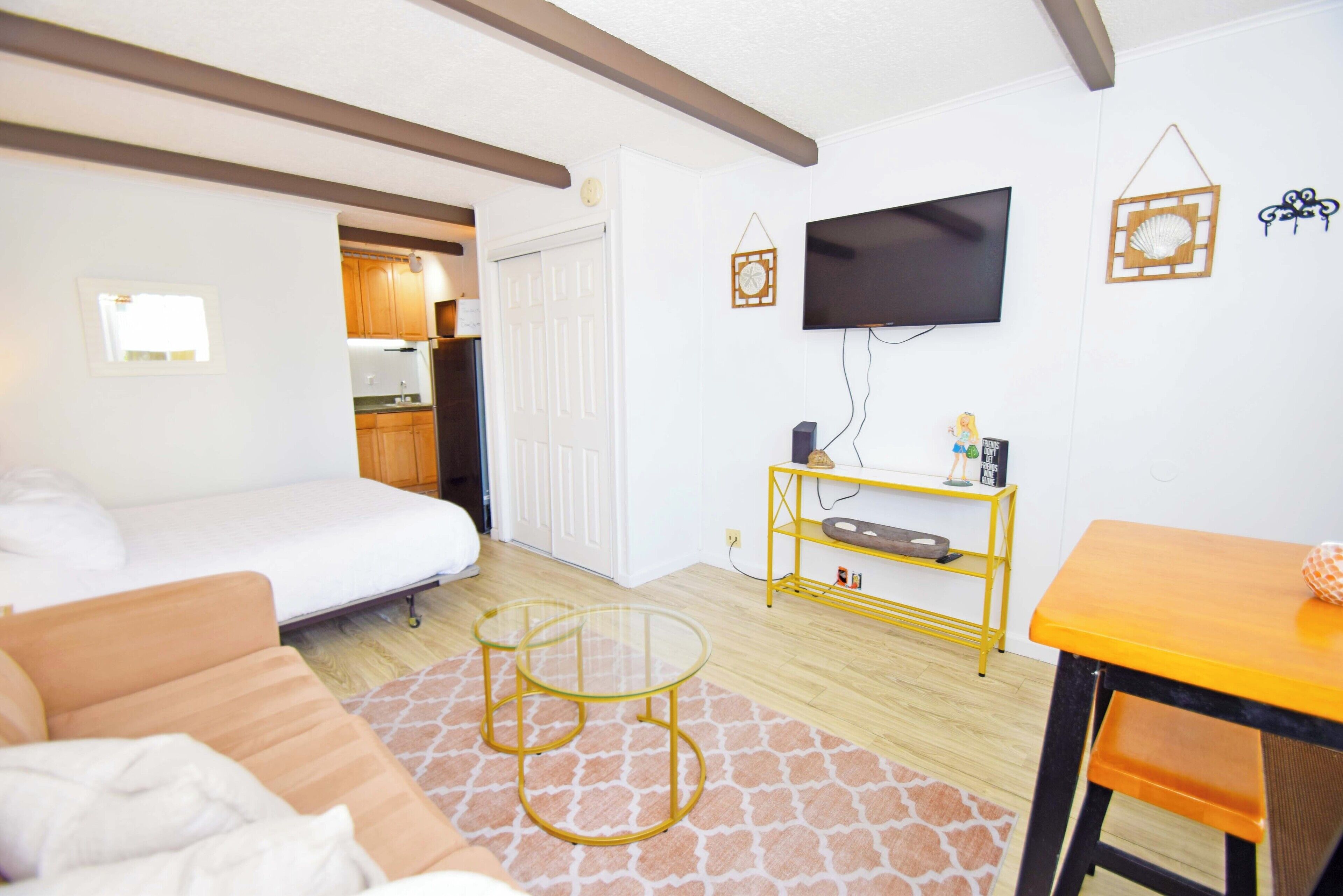 A compact living space with a bed, sofa, small round tables, a TV, and a kitchenette.