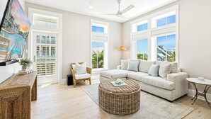 A spacious living room with a large window, a ceiling fan, a wicker ottoman, and a white sofa.