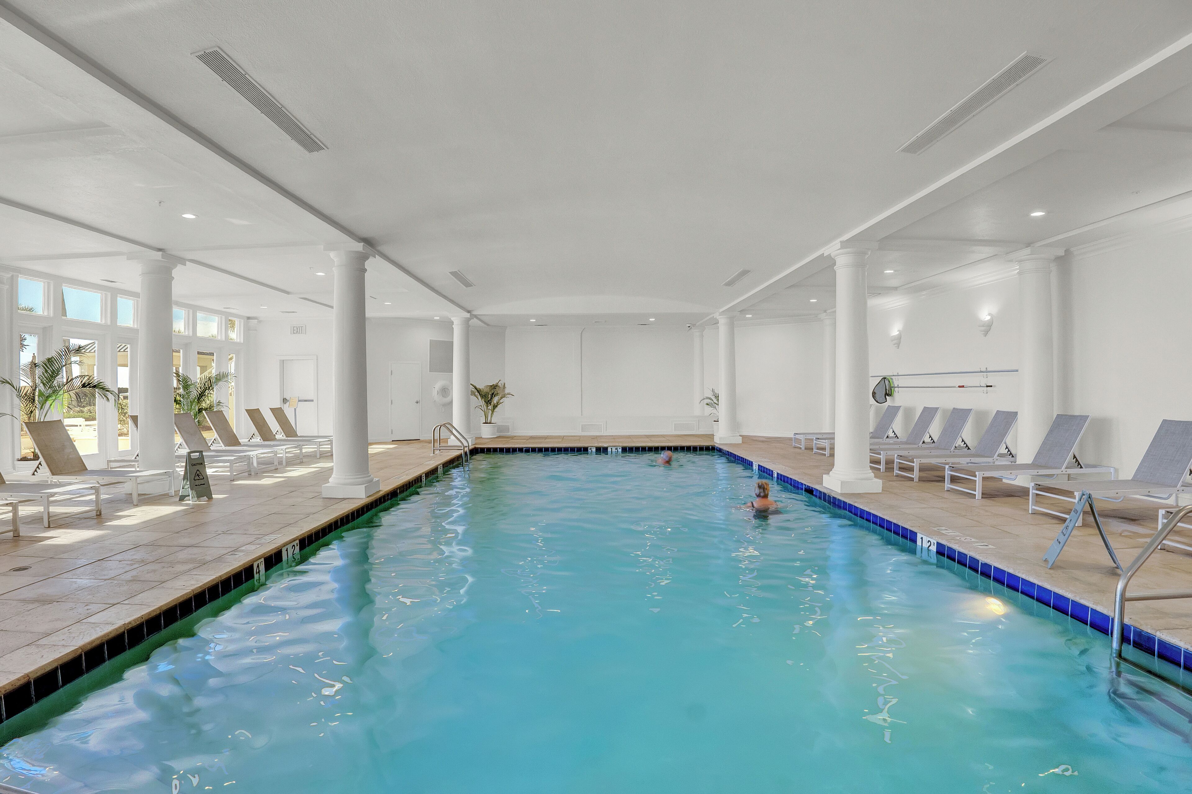 Une piscine intérieure à l’eau claire, entourée de colonnes blanches et de coins ensoleillés aménagés avec des chaises longues.