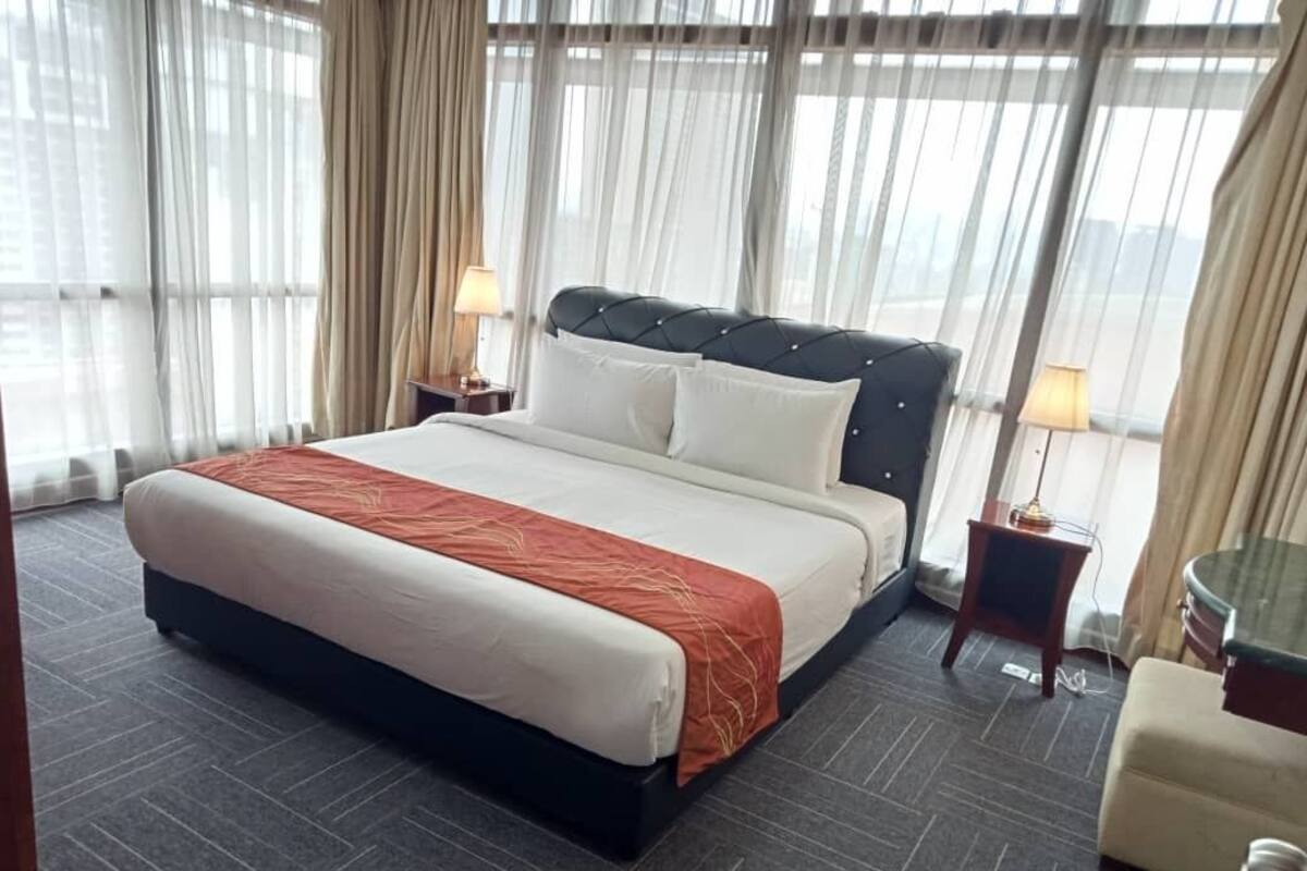 A hotel room with a large bed, two bedside tables, a nightstand, and a view through sheer curtains.
