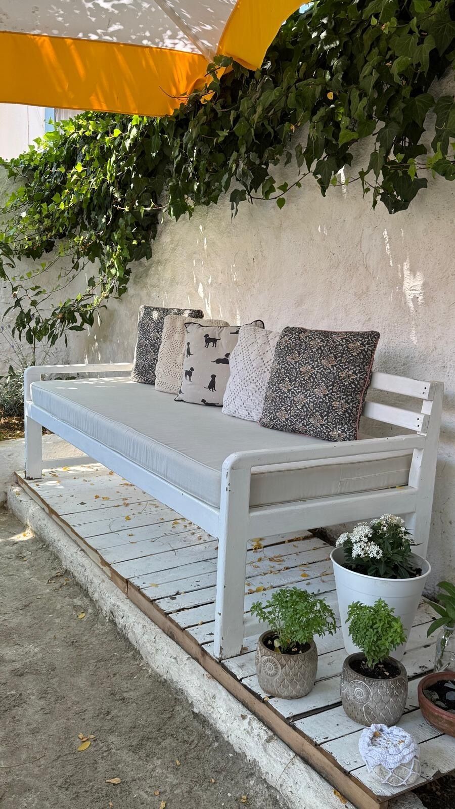 Un banc blanc placé à l’extérieur, agrémenté de coussins à motifs, entouré de plantes en pot et adossé à un mur recouvert de lierre.