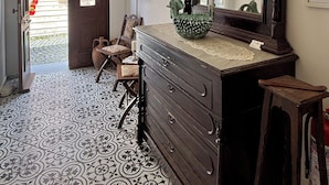 A room with a chandelier, a large mirror, a wooden dresser, patterned floor tiles, and a door leading to an outdoor area. - Vasiliki Konak (Bozcaada)