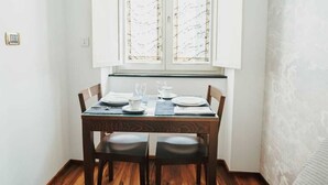 A dining area with a wooden table set for two, a window with curtains, and a sofa in the foreground.