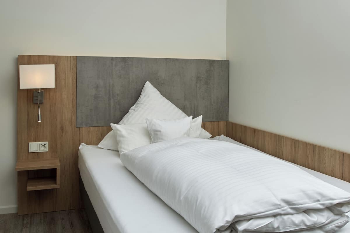 A modern hotel room with a neatly made bed, a wooden nightstand with a lamp, and a grey headboard.