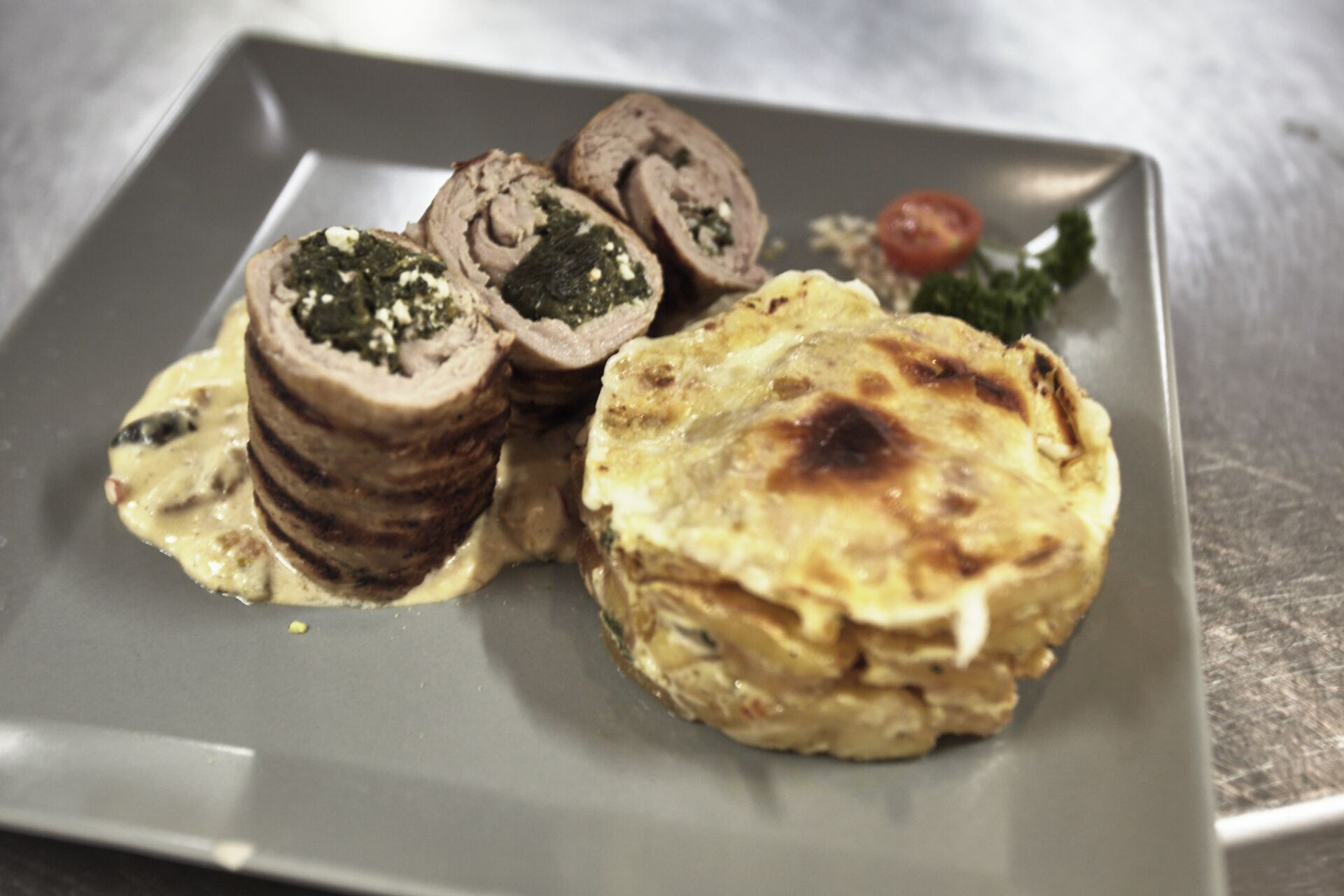 A plate with rolled meat, a baked item, and a small tomato on a gray plate.
