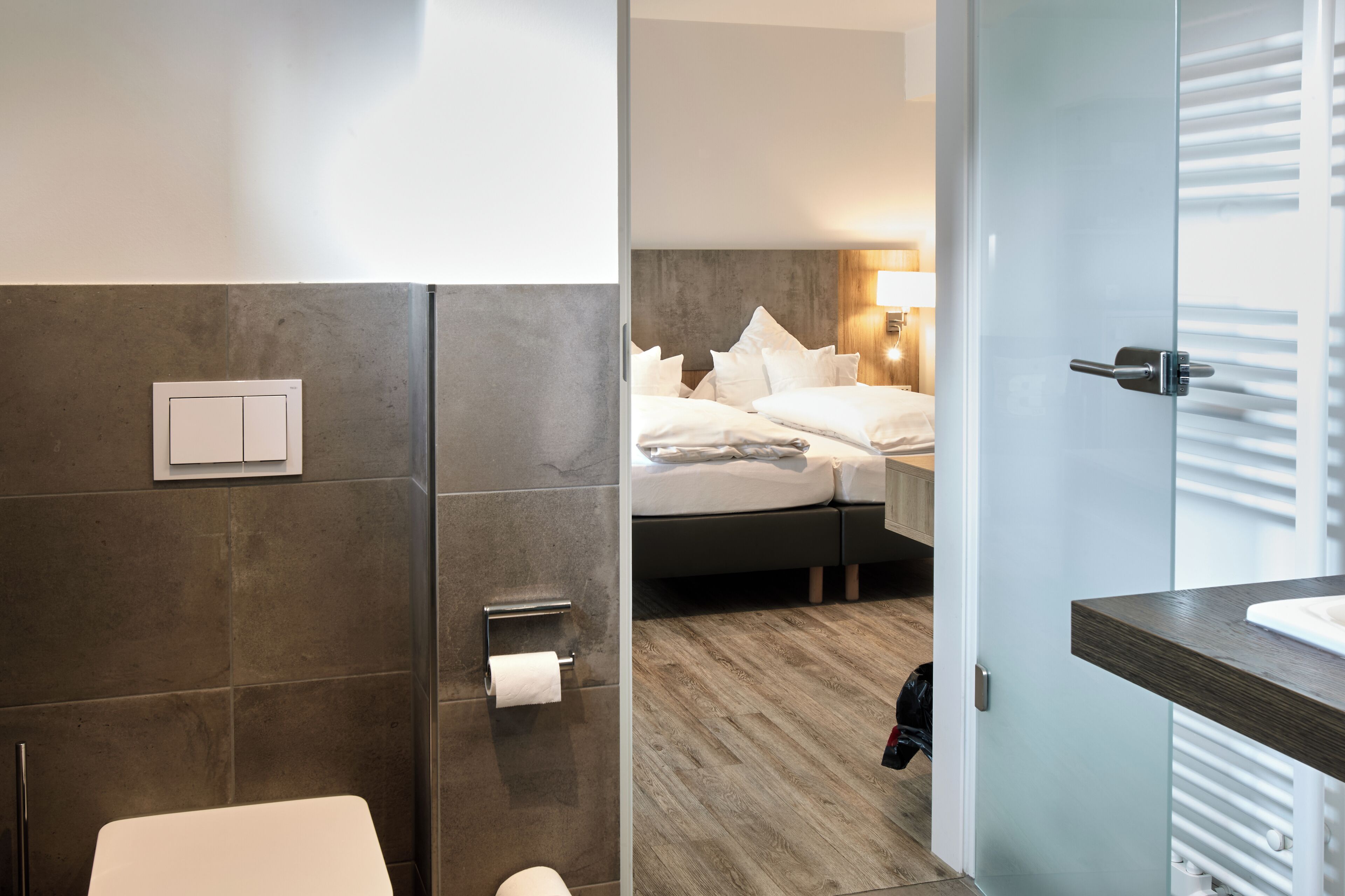 A modern hotel bathroom with a shower, toilet, and a glass-enclosed bedroom with a bed and bedside table.