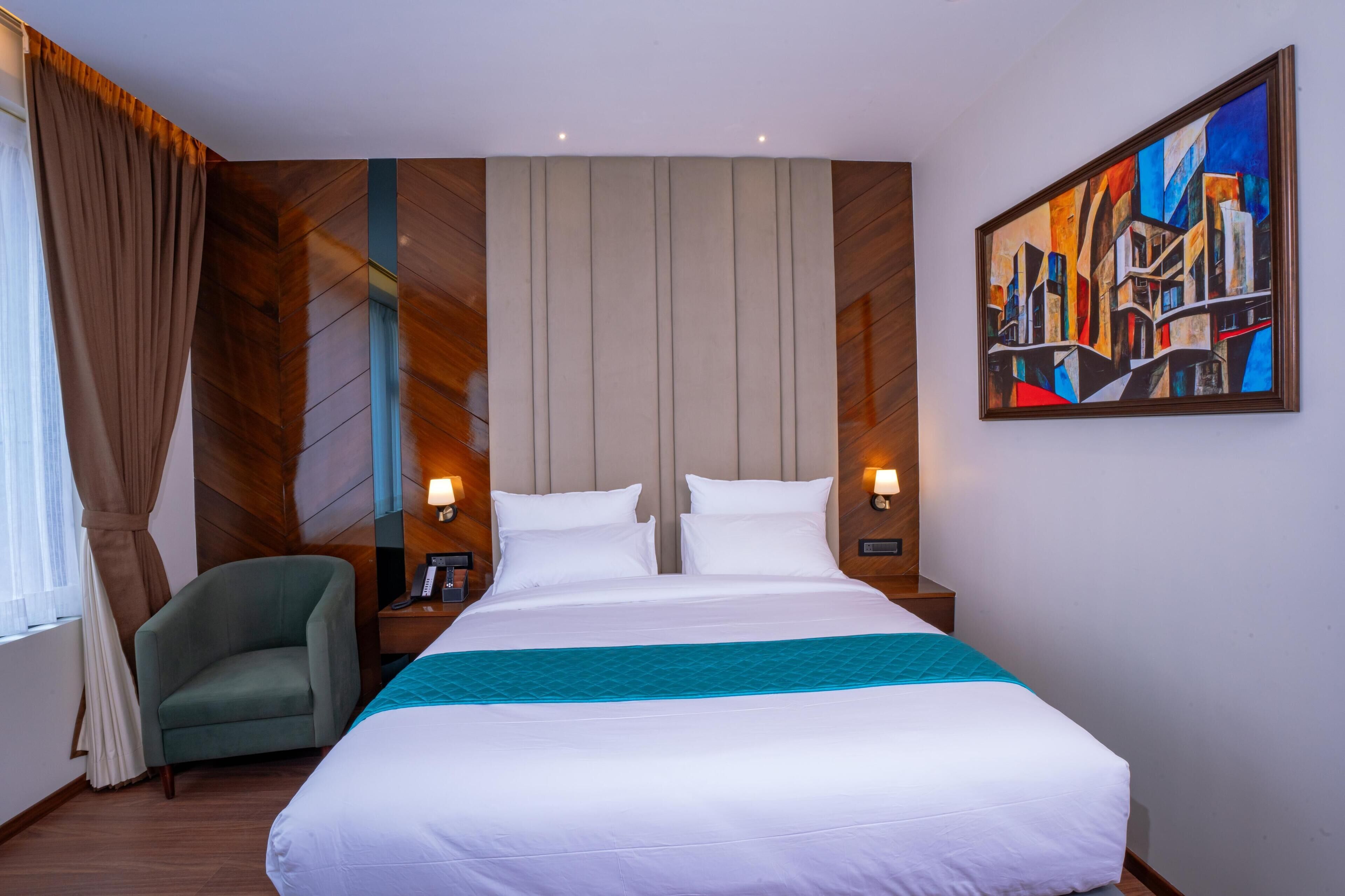 Une chambre d’hôtel avec un grand lit, un fauteuil vert, un tableau au mur et une fenêtre avec des rideaux.