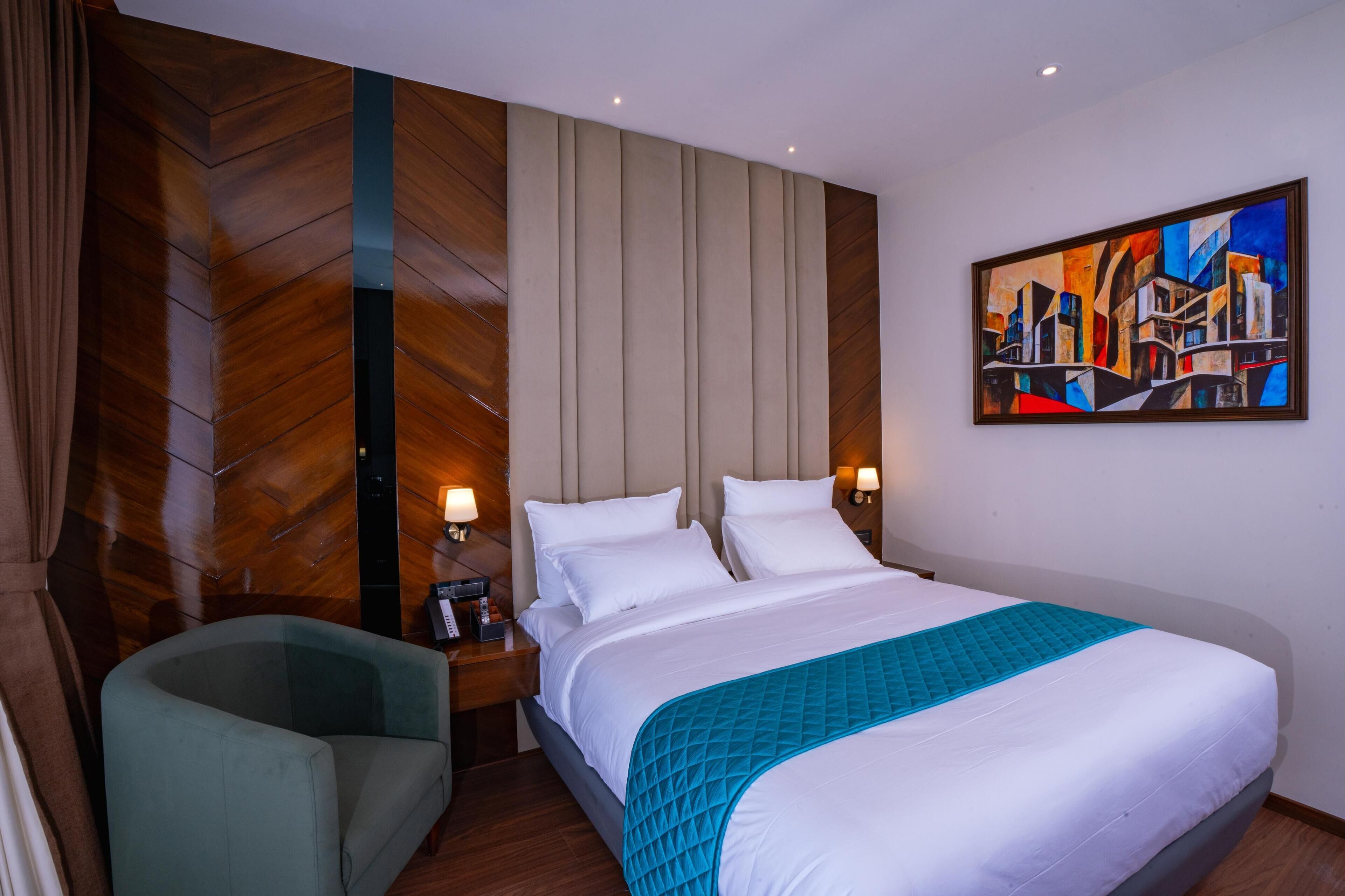 A hotel room with a large bed, a nightstand with a lamp, a chair, and a painting on the wall.