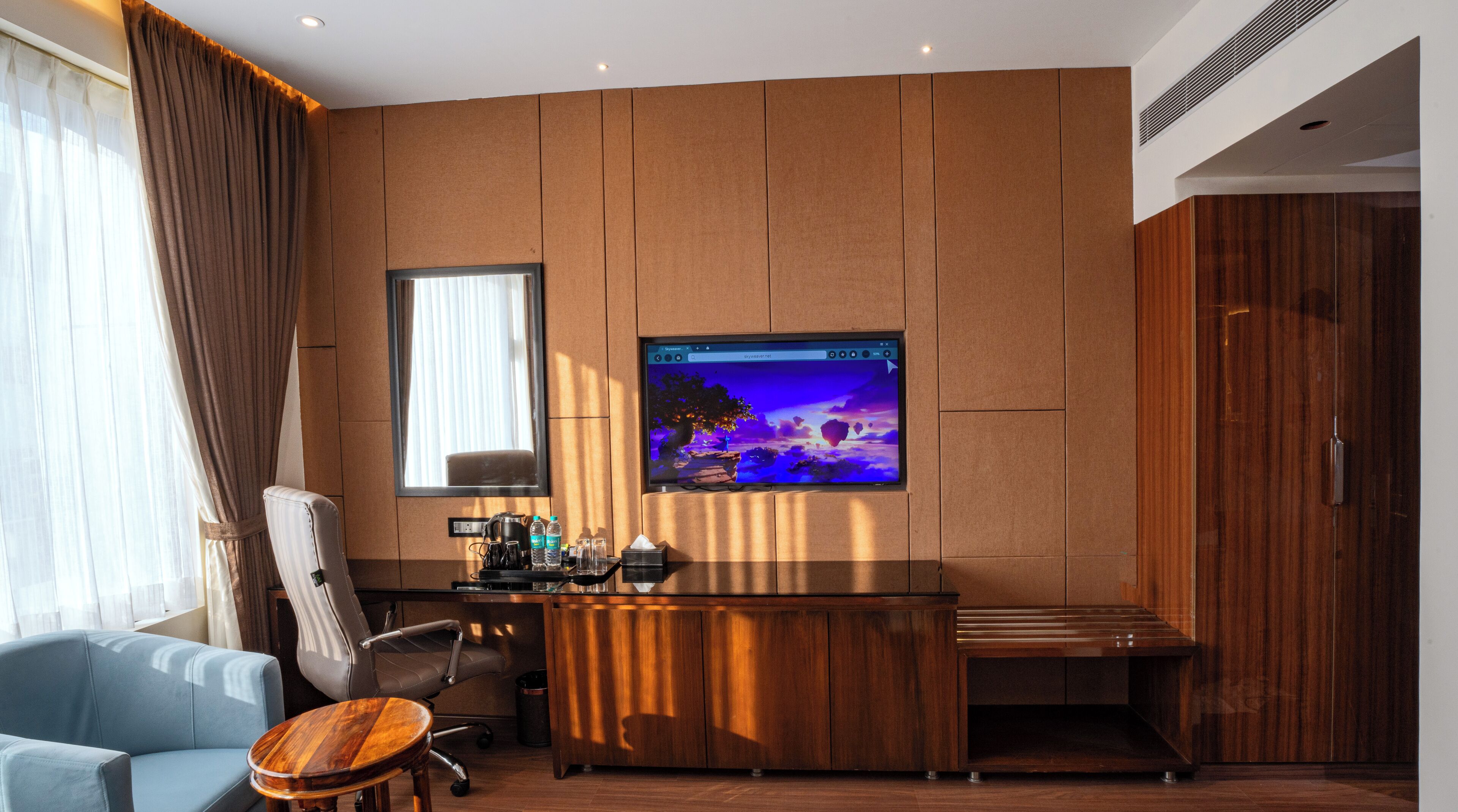 A modern hotel room with a wooden panel wall, a large flat-screen TV, a desk with a chair, and a sofa.