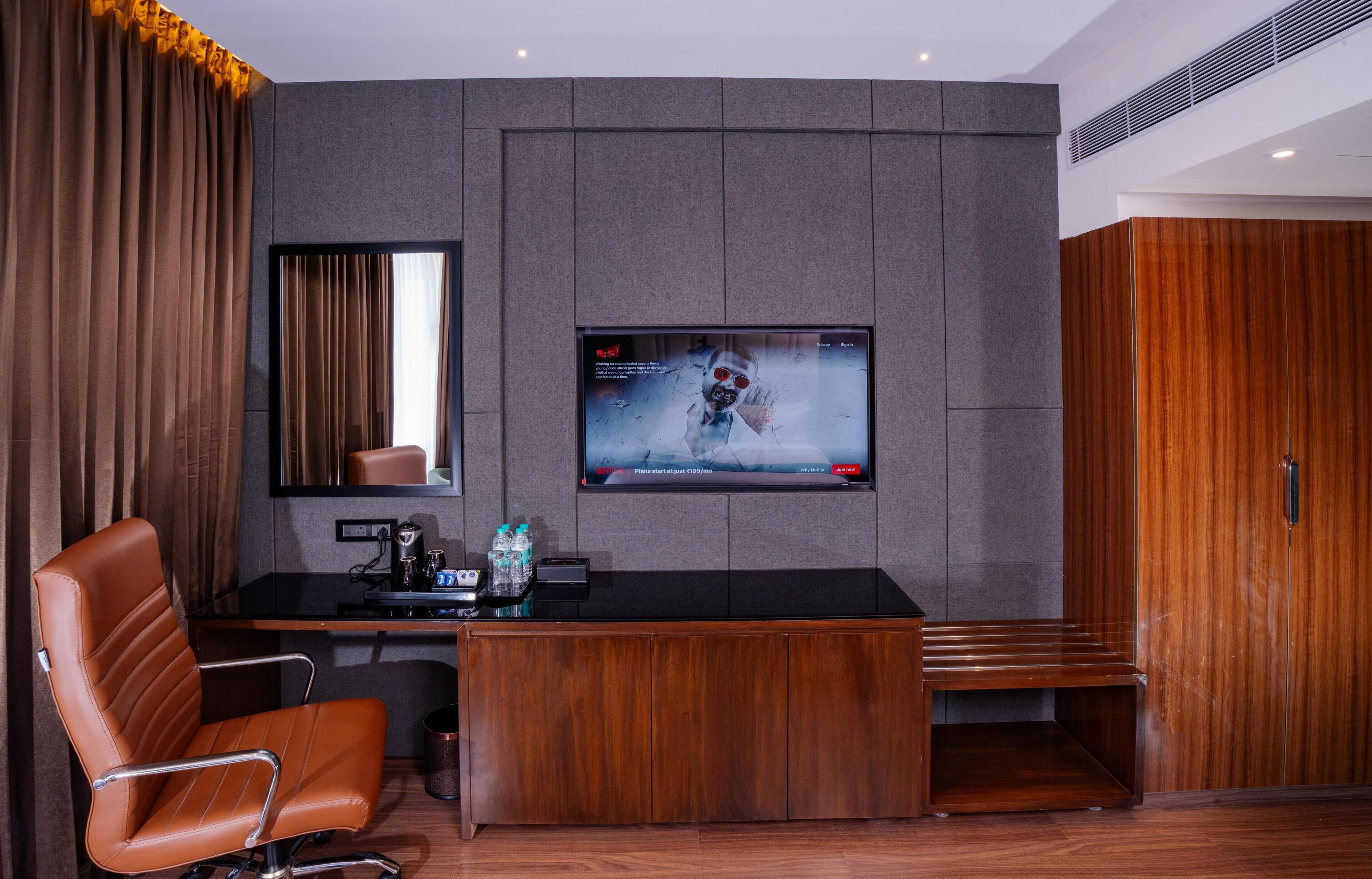 Une chambre d’hôtel moderne avec un bureau en bois, un fauteuil en cuir, un grand écran de télévision et une armoire.
