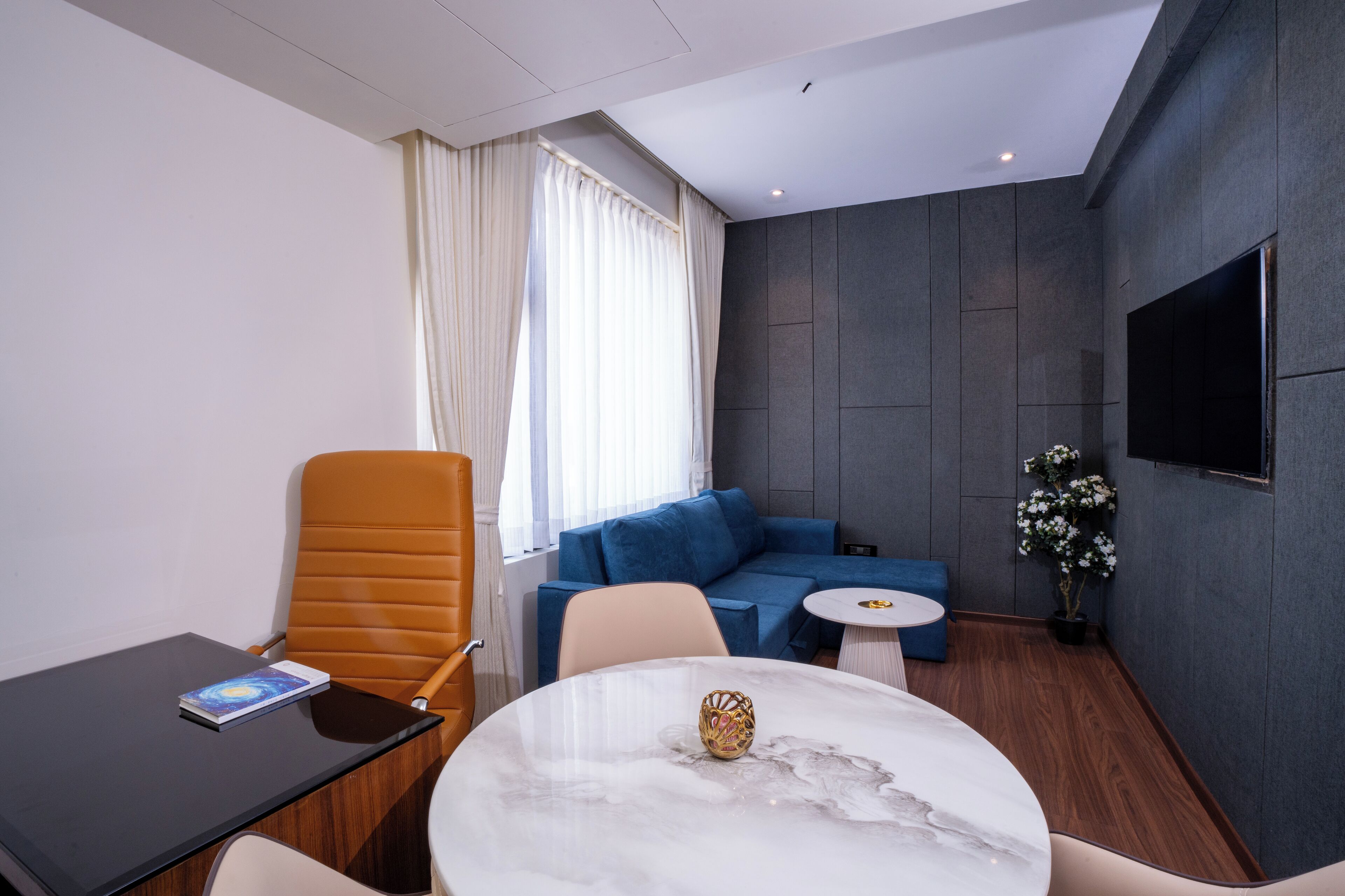Quarto de hotel moderno com uma mesa de mármore, um sofá azul, uma cadeira laranja e uma pequena mesa redonda, com uma TV fixada na parede.