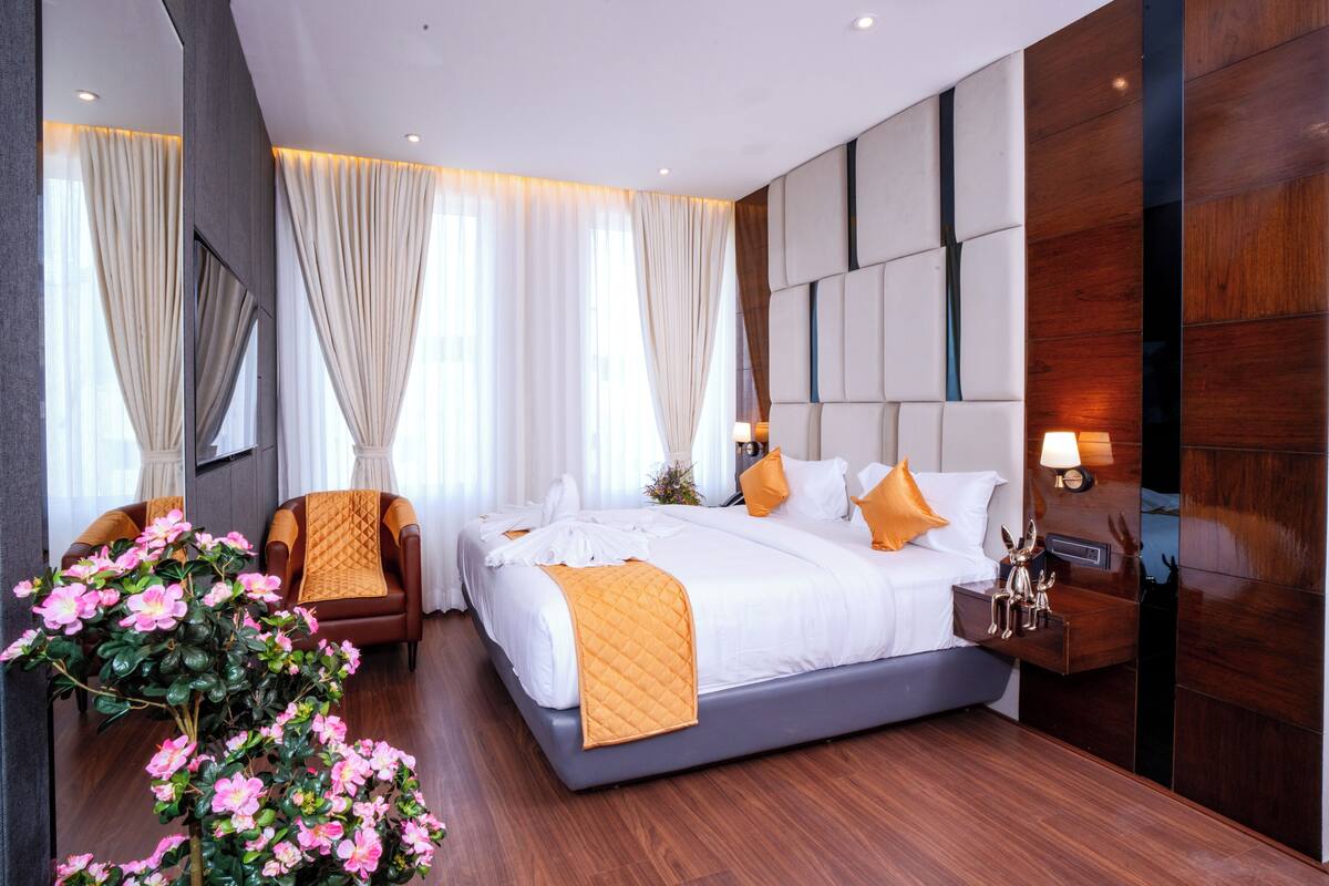 A modern hotel room with a large bed, a seating area with a chair, a nightstand, and a decorative plant.