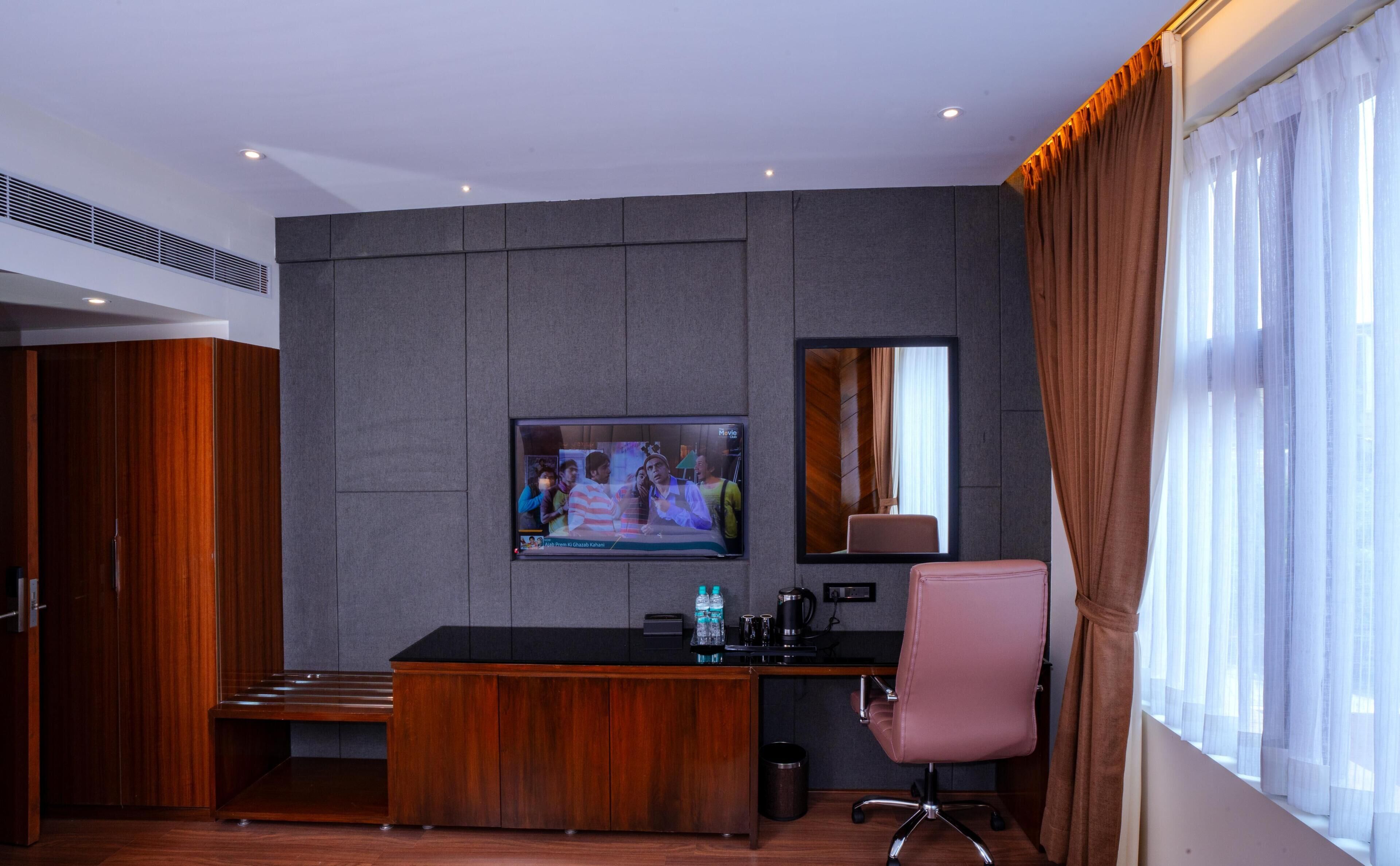 A modern hotel room with a wooden desk, a television mounted on the wall, a large mirror, and a window with curtains.