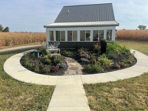 A house with a metal roof, surrounded by a garden with various plants and flowers, and a pathway leading to the entrance.
