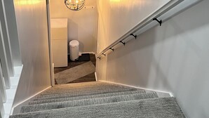 Un escalier étroit avec un palier recouvert de moquette, un mur blanc, une petite fenêtre avec une horloge et une porte en haut.