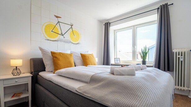 A modern bedroom with a large bed, a bedside table with a lamp, a window with curtains, and a wall-mounted bicycle art piece. - Cozy apartment fully equipped J&K Apartments (Kelheim)