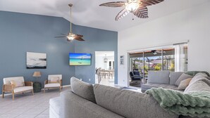 Un salon avec un canapé gris, deux fauteuils, un ventilateur de plafond et un téléviseur fixé au mur.