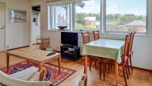 Un salon avec une table en bois, des chaises, un téléviseur et une grande fenêtre donnant sur des maisons.