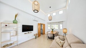 Un salon spacieux avec un canapé beige, un téléviseur fixé au mur, un coin repas comprenant une table et des chaises, et une grande fenêtre laissant entrer la lumière naturelle.