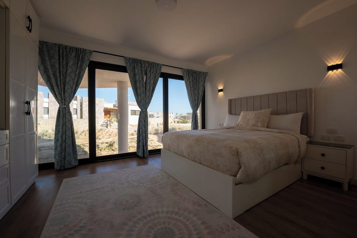 A bedroom with a large bed, a nightstand, a wall-mounted light, and a view of the outside.