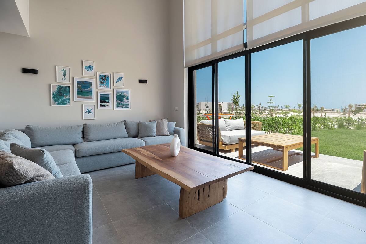 A modern living room with a grey sofa, a wooden coffee table, and a large window offering a view of the outdoors.