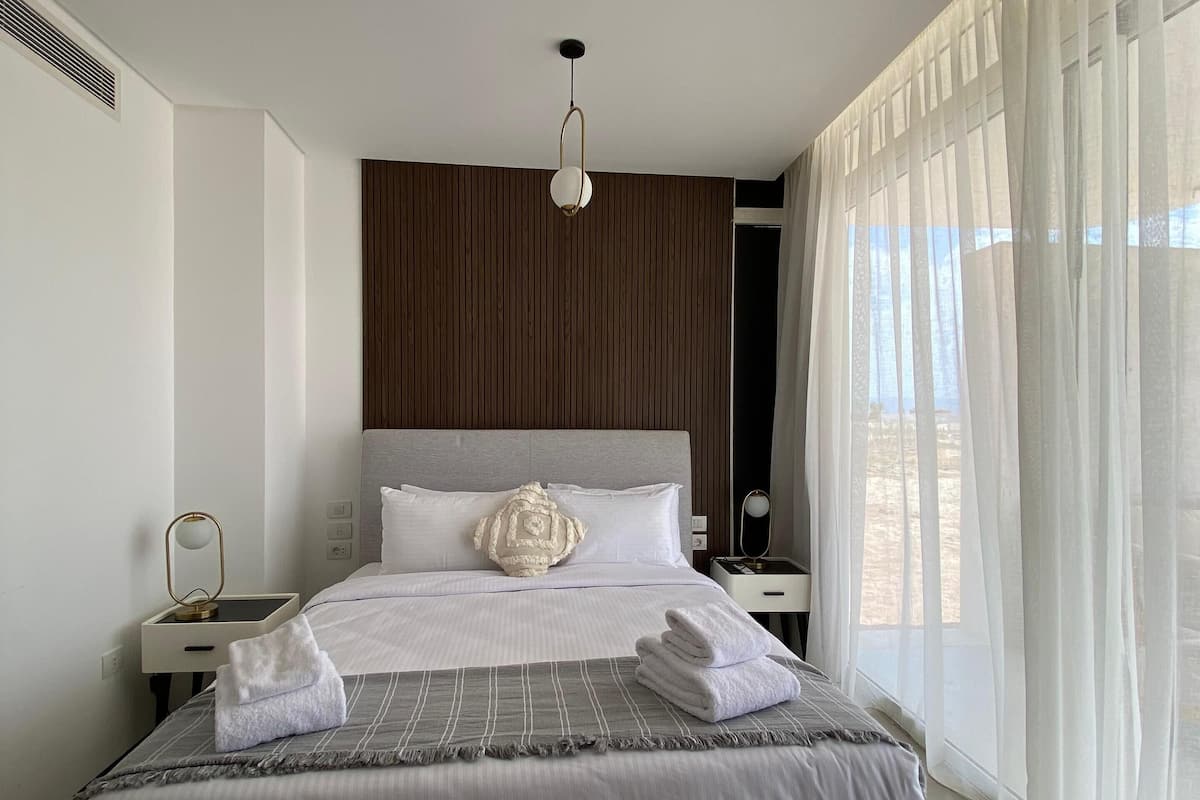 A modern hotel room with a large bed, a bedside table, a nightstand, a hanging light fixture, and sheer curtains.