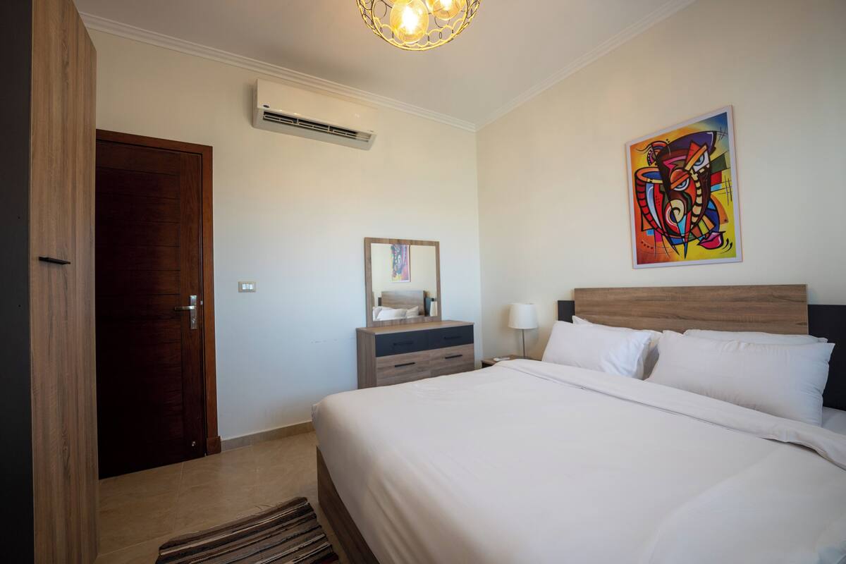 A hotel room with a wooden door, a bed with white linens, a nightstand with a lamp, a dresser, and a colorful abstract painting on the wall.