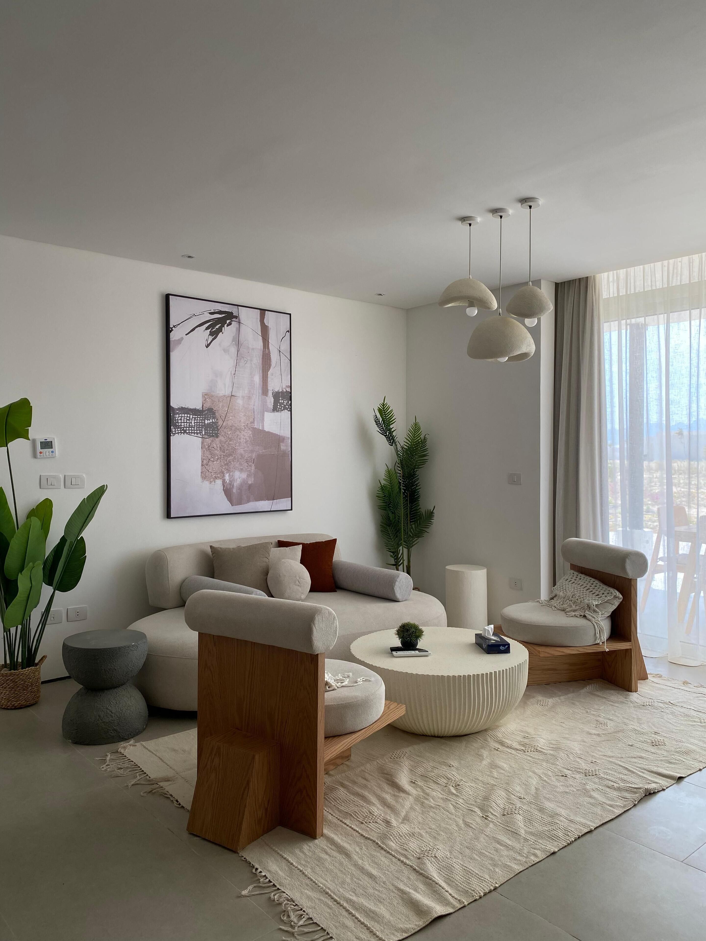 A modern living room with a sofa, coffee table, and side table. There is a large window with curtains, a plant, and a painting on the wall.