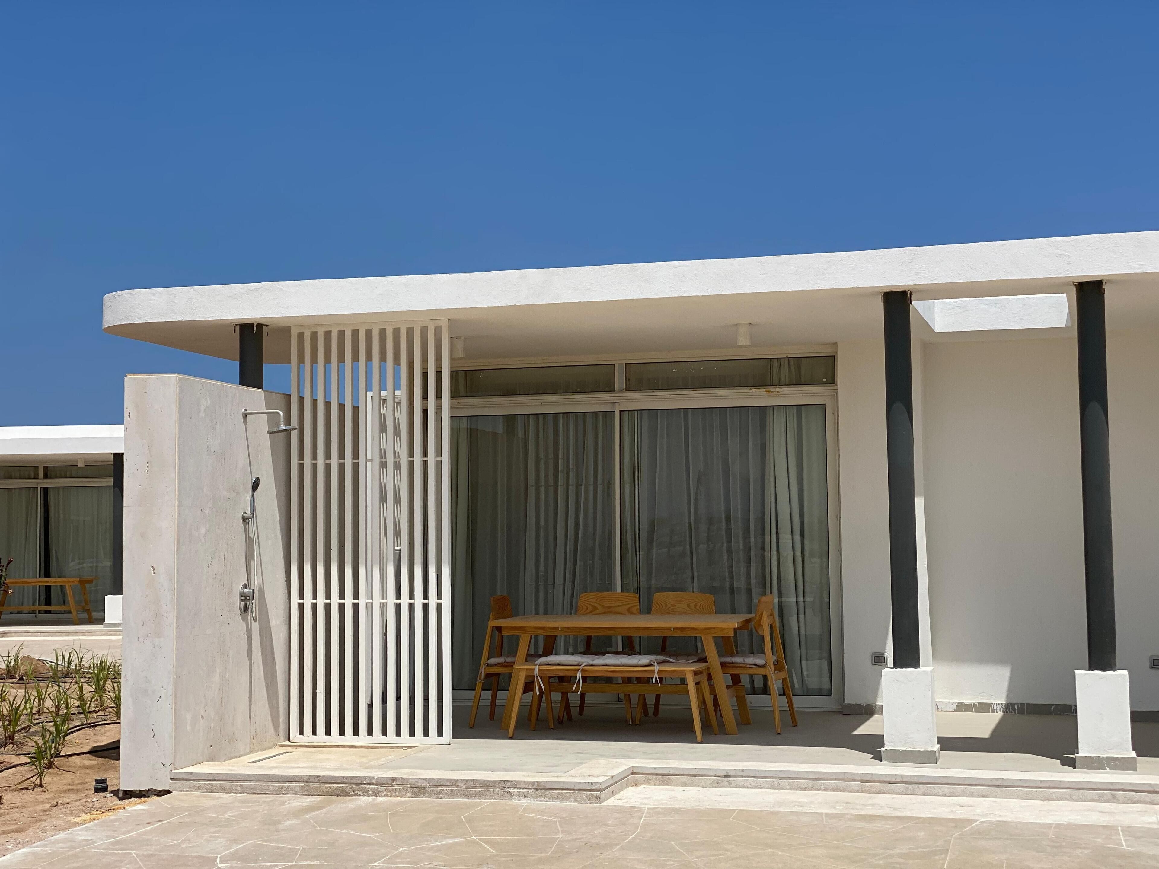 A modern outdoor patio area with a wooden table and chairs, a white gate, and a glass door leading to a covered space.