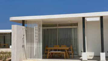 A modern outdoor patio area with a wooden table and chairs, a white gate, and a glass door leading to a covered space.