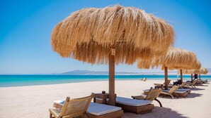 A beach with straw umbrellas and lounge chairs, a clear blue sky, and a calm sea. - Stayr Somabay - Bay life home rentals (Soma Bay)