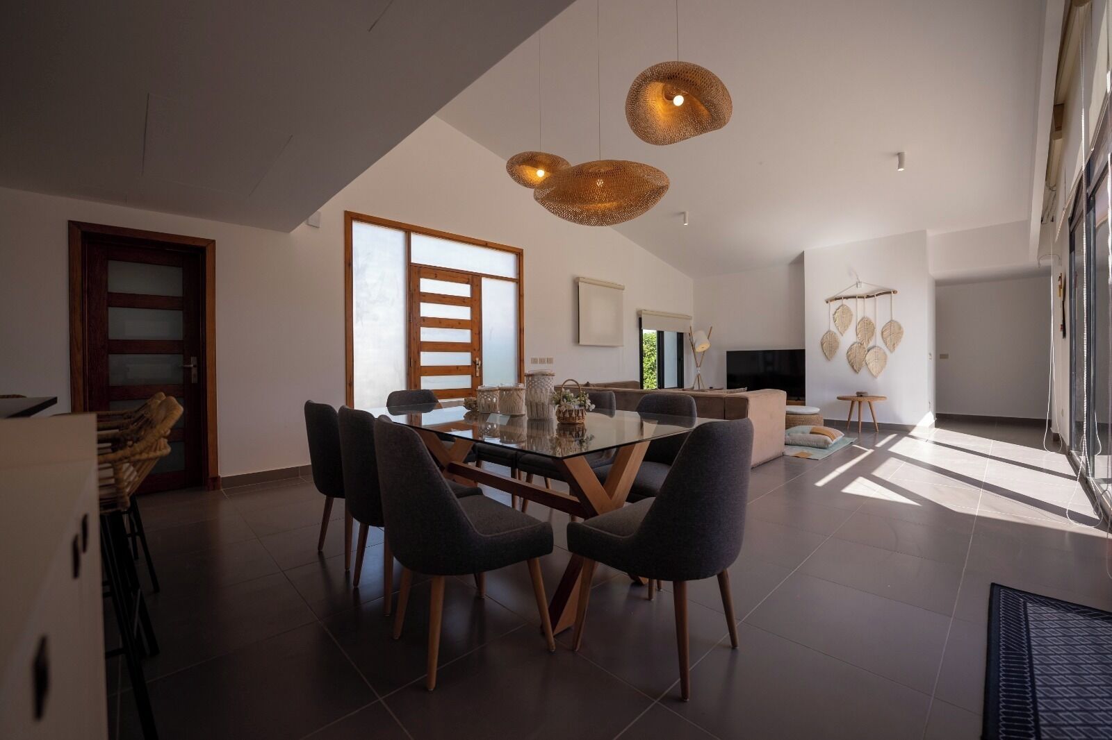 A modern dining area with a glass table and dark chairs, a kitchen in the background, and a large window providing natural light.