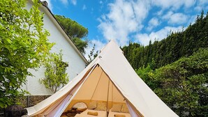Een tent met twee bedden en een kleine tafel, opgesteld in een tuin met bomen en een wit gebouw op de achtergrond.
